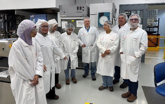A group of participants wearing white lab coats, hairnets and safety gear stand together in a laboratory while an instructor speaks to them. They appear to be receiving a tour or briefing amid lab equipment and industrial workstations.